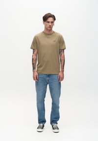 Tricou verde măsliniu cu mâneci scurte și un mic logo negru, asortat cu blugi deschis la culoare și adidași negri cu un model alb și negru.