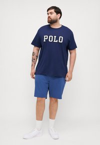 T-shirt azul marinho com o logótipo "POLO" em branco, combinado com calções azuis. Usado com ténis brancos e meias de cano curto, apresentando um corte descontraído.