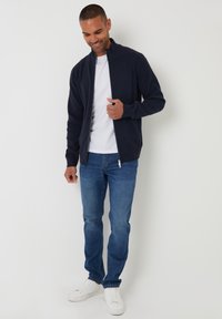 Giacca zip-up blu realizzata in tessuto morbido, indossata sopra una maglietta bianca, abbinata a jeans blu e sneaker bianche. Design semplice con colletto.