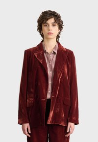Czerwony aksamitny żakiet z klapami, podwójnym zapięciem i dwoma przednimi kieszeniami. Noszony na koszuli w kratę. Gładka tekstura, błyszcząca powierzchnia.