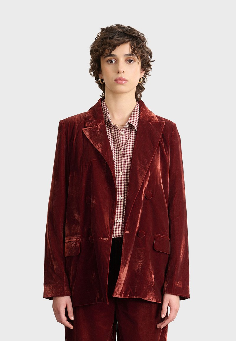 Czerwony aksamitny żakiet z klapami, podwójnym zapięciem i dwoma przednimi kieszeniami. Noszony na koszuli w kratę. Gładka tekstura, błyszcząca powierzchnia.