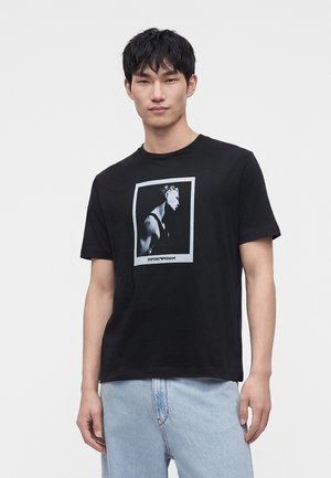 Giovane uomo con indosso una T-shirt nera con una grafica in scala di grigi del profilo di una persona e jeans azzurri su uno sfondo uniforme.