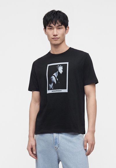 Νέος άνδρας που φοράει μαύρο T-shirt με γκριτονική γραφική απεικόνιση προφίλ ανθρώπου και ανοιχτό μπλε τζιν σε απλό φόντο.