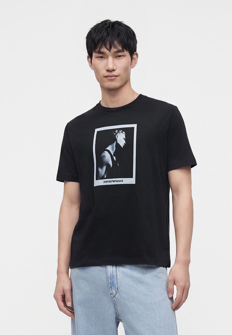Νέος άνδρας που φοράει μαύρο T-shirt με γκριτονική γραφική απεικόνιση προφίλ ανθρώπου και ανοιχτό μπλε τζιν σε απλό φόντο.
