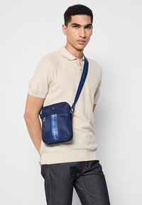 Laivastonsininen olkalaukku, jossa on sinisillä raidoilla varustettu logokoriste, vetoketjullinen tasku ja säädettävä hihna, valmistettu kestävistä kankaista.