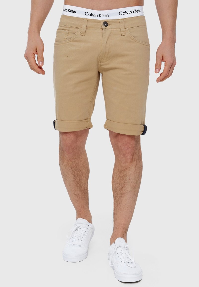 Homme portant un short beige retroussé, un sous-vêtement blanc avec ceinture Calvin Klein, et des baskets blanches, debout devant un fond uni.