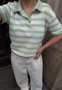 Cilvēks, kurš valkā zaļu un baltu svītrainu adītu polo kreklu ar īsām piedurknēm un baltas bikses, stāv ar vienu roku uz gurna pie akmens sienas.