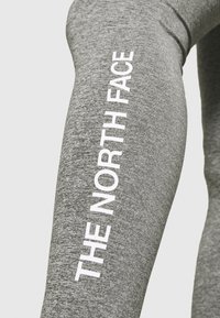 Šedé legíny vyrobené z texturované tkaniny s bílým vertikálním potiskem "THE NORTH FACE" podél vnější strany stehna.