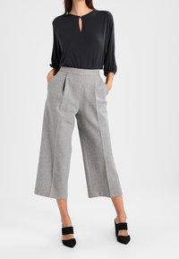 Černá blůza s výstřihem ve tvaru klíčové dírky a dlouhými rukávy, v kombinaci s šedými kalhotami s vysokým pasem a širokými nohavicemi. Celkový vzhled doplňují černé lodičky.