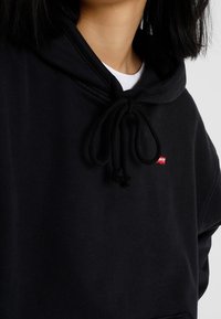 Sweat à capuche en coton noir avec un col à cordon de serrage, une poche frontale et une petite étiquette rouge Levi's sur la poitrine. Texture douce avec une coupe décontractée.