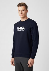 Marineblaues Sweatshirt mit rundem Halsausschnitt und langen Ärmeln, das ein auffälliges weißes "KARL LAGERFELD"-Logo auf der Vorderseite trägt. Glatte Textur.