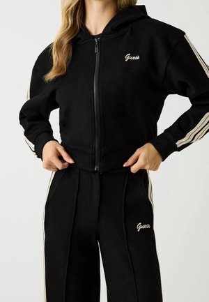 Frau trägt einen schwarzen Zip-Hoodie und passende Hose mit beigen Streifen an Ärmeln und Hosenbeinen, beide mit dem Schriftzug "Guess" in beiger Farbe.