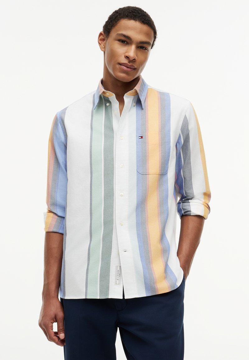 Homme portant une chemise boutonnée à rayures verticales multicolores avec manches retroussées et pantalon foncé, debout avec une main dans la poche.