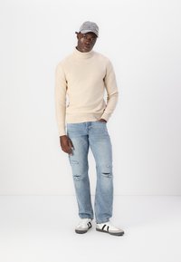 Maglione a collo alto beige, jeans azzurri strappati, e sneakers bianche con strisce nere. Cappellino grigio con accenti colorati.