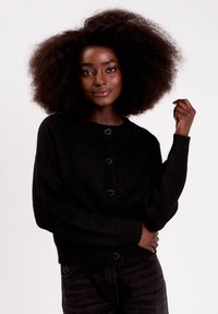 Cardigan noir en tricot avec des boutons en forme de cœur, manches longues et coupe décontractée. Porté sur un jean foncé, sur un fond neutre.