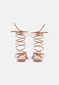 Bruine leren sandalen met spiraalvormige enkelbanden, platte zolen en een minimalistisch ontwerp. Gladde textuur en gestrikte strikken bovenaan elke band.