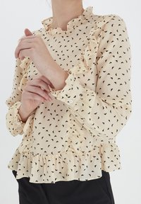 Blusa crema con colletto e polsini a volant, stampa a cuoricini neri e orlo a balze e svasato. Realizzata in tessuto leggero.