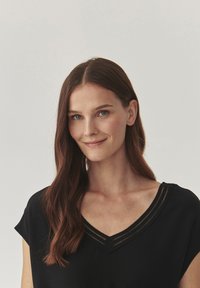 Haut noir à manches courtes, décolleté en V avec une bordure texturée, tissu lisse et éclat subtil. Le modèle a de longs cheveux bruns.
