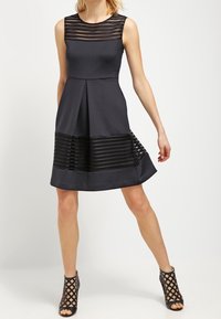 Robe noire sans manches avec un haut en dentelle texturée et un bas plissé et lisse. Elle présente une silhouette ajustée et des accents semi-transparents.