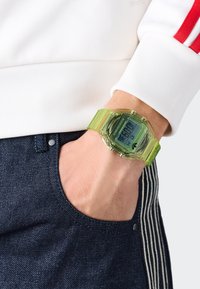 Ceas digital cu un brățară din plastic transparent verde, față rectangulară și display digital negru. Poartă pe un încheietură alături de blugi negri.