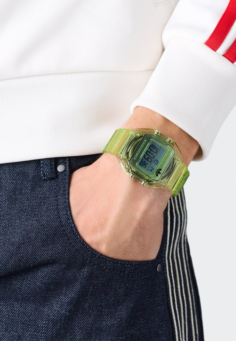 Skaitmeninis laikrodis su skaidriu žaliu plastiko dirželiu, stačiakampiu ciferblatu ir juodu skaitmeniniu ekranu. Nešiojamas ant riešo kartu su tamsiais džinsais.