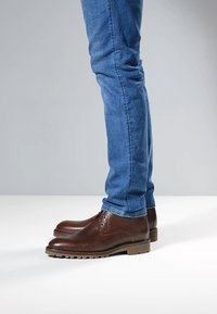 Scarpe in pelle marrone con texture, tacco basso, suola in gomma e cuciture dettagliate, abbinate a jeans blu a taglio slim.