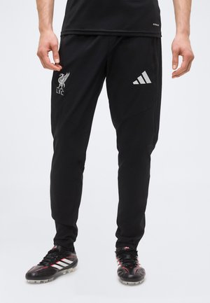 Mann trägt schwarze Liverpool FC Sporthose mit Adidas-Logo, steht, trägt auch schwarze Adidas Fußballschuhe mit weißen Streifen und roten Akzenten.
