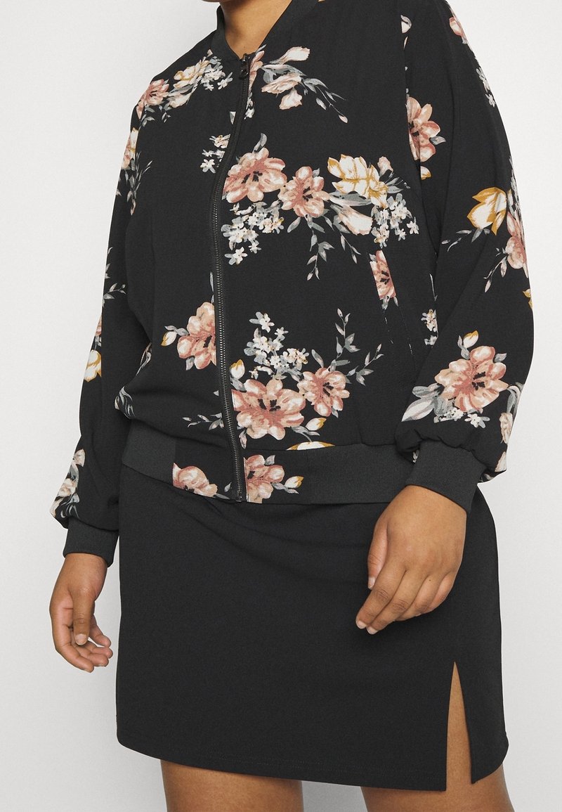 Svart blommig bomberjacka med dragkedja, prydd med rosa och vita blommönster på ärmarna; matchas med en svart minikjol med en sidofåra.