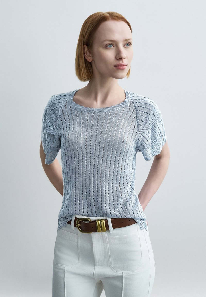 Pull tricot bleu clair à manches courtes, avec rayures verticales et texture côtelée, porté avec un pantalon blanc et une ceinture marron.