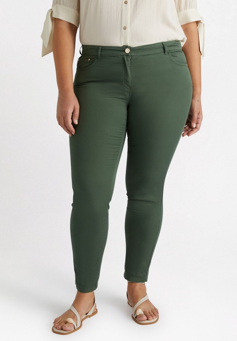 Kvinna som bär mörkgröna höga skinnybyxor, en beige blus med knutade ärmar och beiga sandaler med tåslingor, stående mot vit bakgrund.