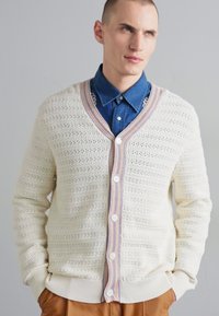 Cremefarbener gestrickter Cardigan mit V-Ausschnitt, Knopfverschluss und gemustertem Saum. Mit Rippbündchen an den Ärmeln und dem Bund, getragen über einem blauen Hemd.