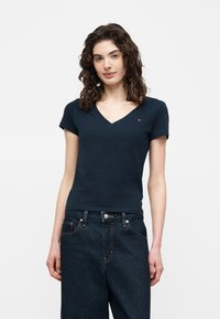 Γυναίκα με σγουρά μαλλιά που φοράει εφαρμοστή σκουρόμπλε V-λαιμόκοψη μπλούζα και ψηλόμεσο σκούρο τζιν σε απλό φόντο.