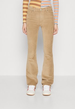 Beige Cord-Hose mit Schlag, getragen mit weißen Turnschuhen und einem mehrfarbig gestreiften Langarmshirt, stehend vor einem einfarbigen Hintergrund.