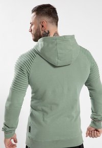 Sudadera verde con acentos acanalados en los hombros, tela texturizada, bolsillo canguro y capucha, con un dobladillo recto y mangas ajustadas.