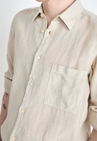 Camicia button-up in lino beige chiaro, con una piccola tasca frontale e maniche lunghe, dotata di bottoni in conchiglia e una finitura rilassata e texture.