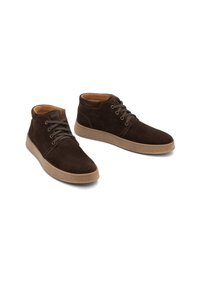 Une paire de bottines en daim marron avec des semelles en caoutchouc beige et des lacets, sur un fond blanc.
