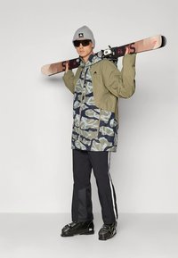 Slidinėjimo apranga su žalia ir jūrine maskuotės rašto striuke, juodos slidinėjimo kelnės su baltais dryžiais ir juodi slidinėjimo batai, laikantys slides.