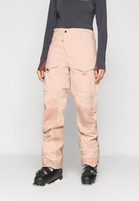 Calças de neve cor-de-rosa com um material à prova d'água, bolsos laterais tipo cargo e detalhes em zíper. Usadas com botas de esqui pretas.