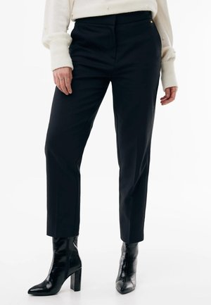 Kvinde iført sorte, skræddersyede bukser til anklerne, hvid sweater med perle detaljer og sorte højhælede ankelstøvler.