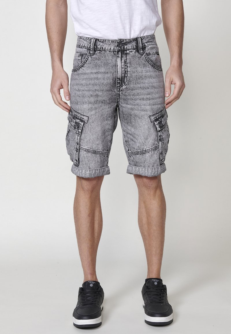 Shorts cargo en denim gris avec ourlets roulés, deux poches avant et poches latérales à rabat. Portés avec des chaussures de sport noires.