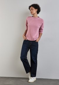 Rosa Fleece-Pullover mit langen Ärmeln, kombiniert mit dunkelblauen Jeans. Die Person trägt weiße Sneakers und steht vor einem neutralen Hintergrund.