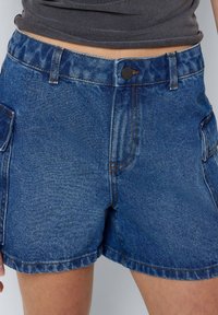 Džínové šortky v středně modré barvě s uvolněným střihem. Mají zapínání na knoflík, mírné obarvení a hluboké boční kapsy.