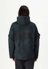 Green and black tie-dye jacket with a hood, featuring a large black print "BOILER ROOM" on the back and multiple utility pockets.