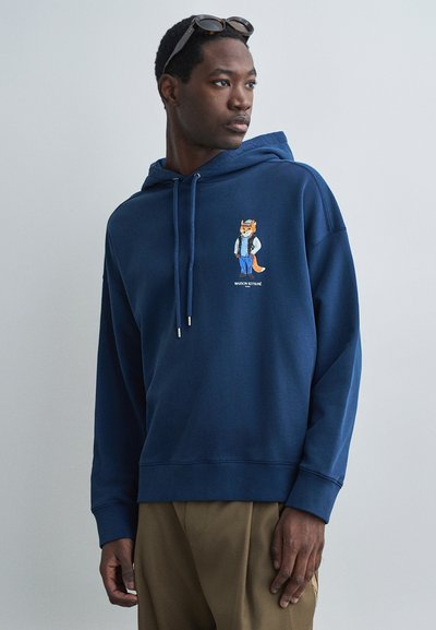 Kapuzenpullover in Marineblau mit einem Cartoon-Fuchs-Design auf der Brust, mit Kordelzug und gerippten Bündchen. Trägt ihn mit beigen Hosen.