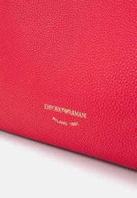 Czerwona torebka skórzana o fakturze z złotym wytłoczonym logo, które brzmi "EMPORIO ARMANI MILANO 1981."