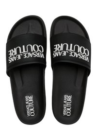 Des sandales noires à glissière en caoutchouc texturé, avec une large bride portant l'inscription "VERSACE JEANS COUTURE" en blanc. Design à semelle plate.