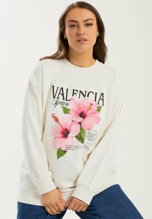 Vrouw draagt een witte sweatshirt met roze hibiscusbloemen en de tekst "Valencia Spain 1987", gecombineerd met een blauwe spijkerbroek.