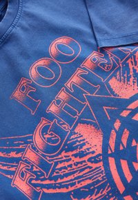 Modra bombažna majica z velikim roza grafičnim napisom "FOO FIGHTER" in okroglimi ter črtastimi vzorci v dizajnu.