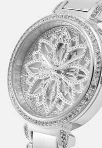 Sølv Guess klokke med en blomsterprydet urskive, krystallakkenter og et skinnende metallarmbånd. Har en gjennomsiktig innpakning og intrikate detaljer.