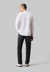 Camicia bianca a maniche lunghe con colletto button-down, abbinata a jeans grigi scuri. La camicia ha un tessuto testurizzato e un'orlo leggermente curvato.